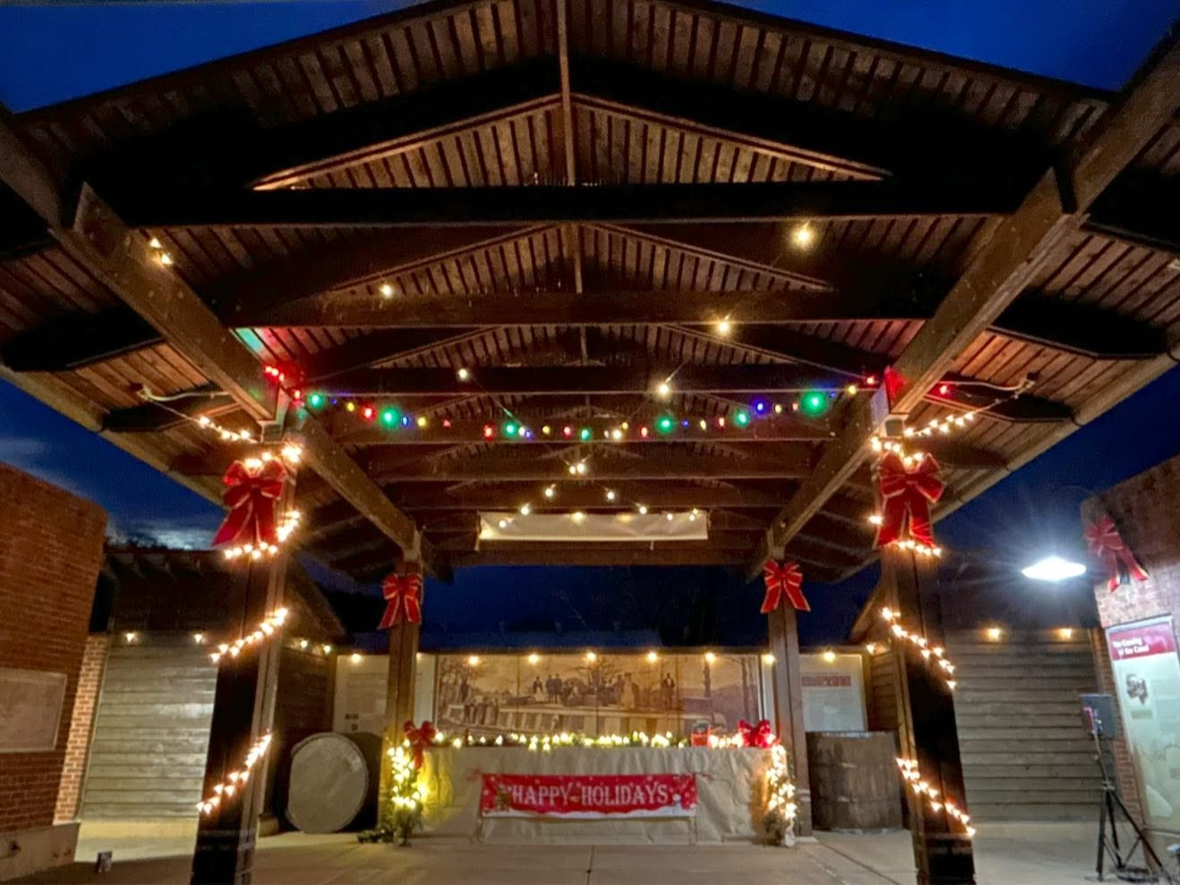 The wooden Pavilion strung with warm lights and red bows, with a Happy Holidays banner