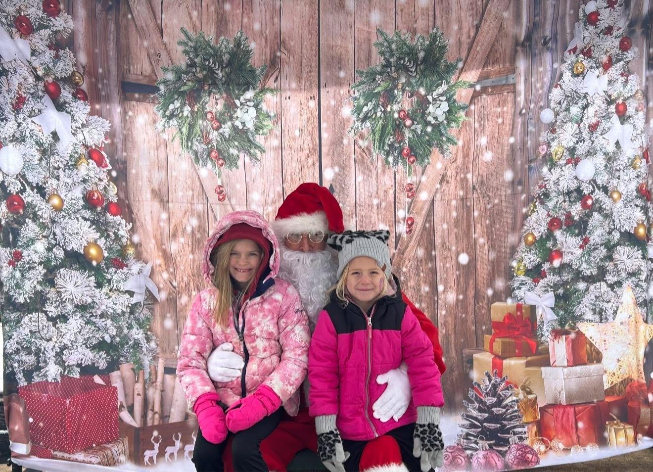Two smiling girls in pink winter coats sitting with Santa in front of a holiday backdrop