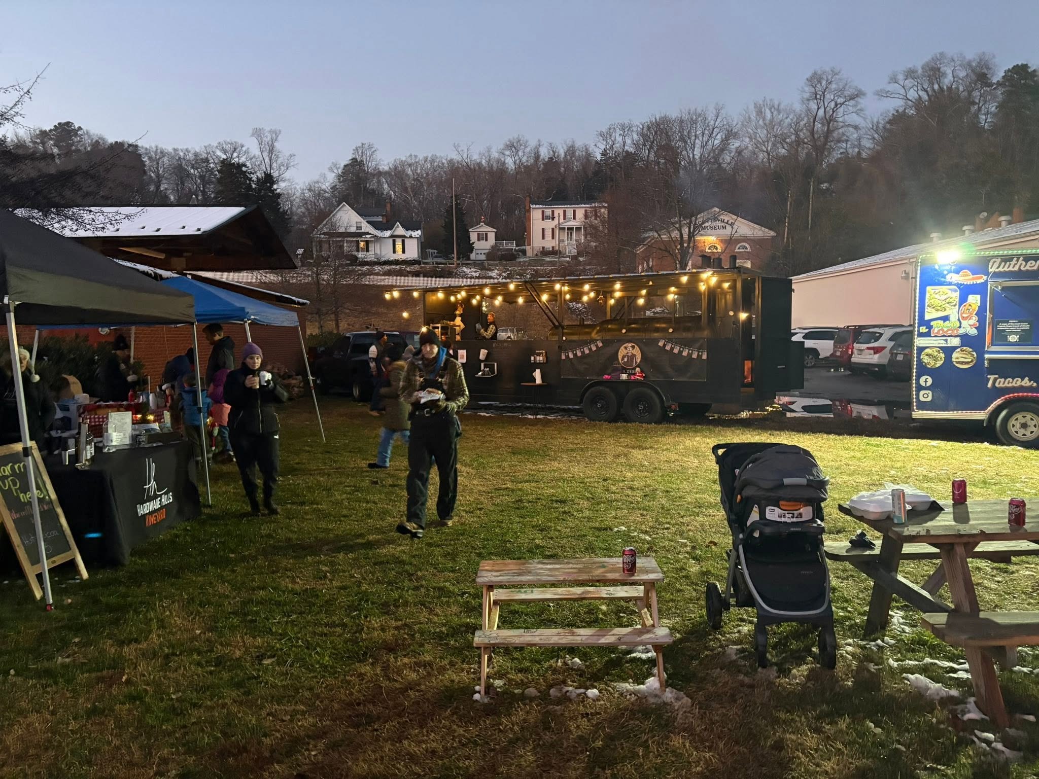 Food trucks, a lit smoker trailer and vendor tents on the lawn at Canal Basin Square at dusk